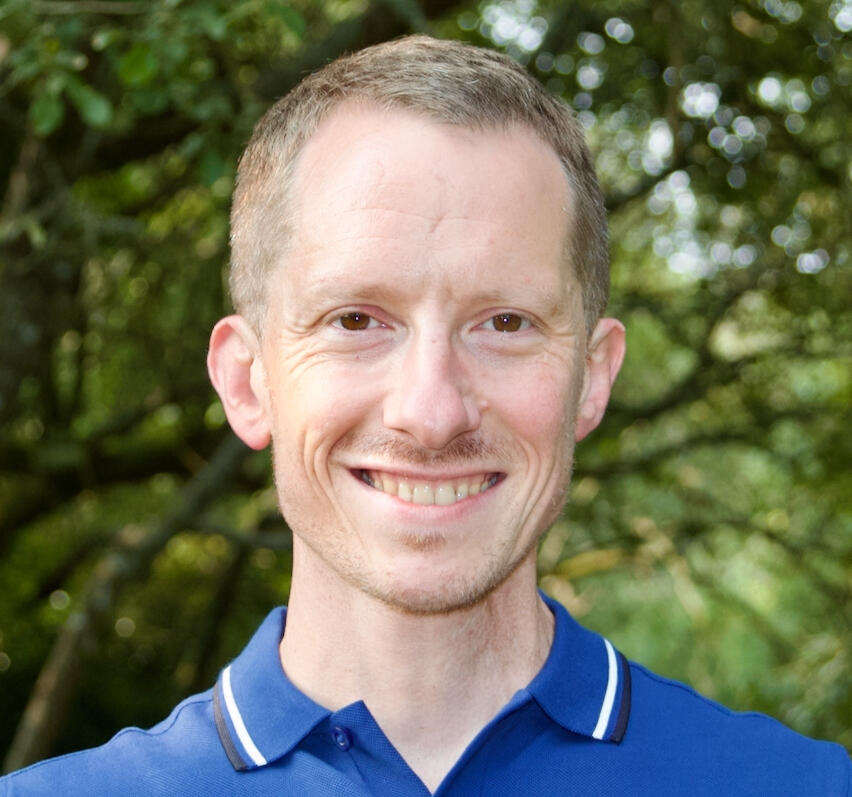 Picture of Tom Burns smiling in outdoor setting, with blue shirt, collar visible and trees in background.